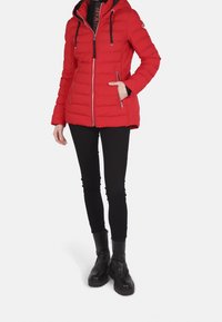 Chaqueta acolchada roja con capucha, cremallera frontal y bolsillos laterales. Presenta detalles en negro y un diseño acolchado. Se lleva con leggings negros y botas.