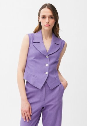 Joven mujer con chaleco sin mangas lavanda con botones plateados y pantalones lavanda a juego, posando contra un fondo blanco liso.