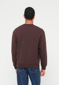 Hnědá mikina s kulatým výstřihem, kartáčovaná textura s žebrovanými manžetami a lemem, kombinovaná s modrými džínami z denimu. Jednoduchý design bez viditelných log nebo vzorů.