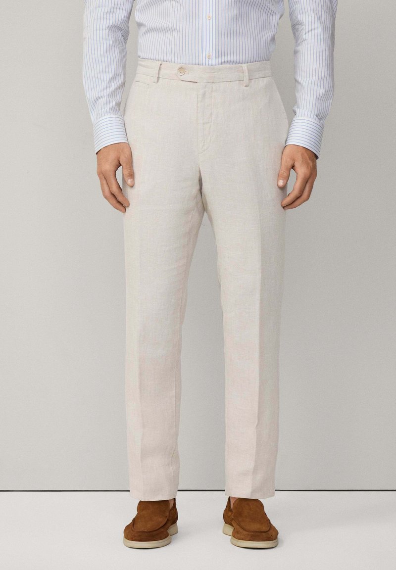 Homme portant un pantalon en lin beige, une chemise rayée bleue et blanche, et des mocassins en suède marron, debout devant un fond uni clair.