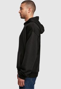 Svart hettegenser med kengurulomme, ribbestrikkede mansjetter og kant; laget av mykt stoff; vist på en modell i sideprofil.