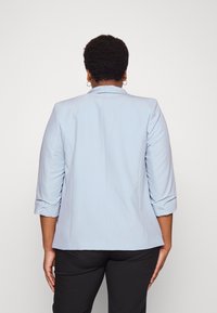 Person with short curly hair wearing a light blue blazer with rolled-up sleeves and black pants, standing facing away on a plain background.