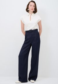 Blouse blanche à manches courtes avec détail en trou de serrure, associée à un pantalon large taille haute bleu marine et une ceinture coordonnée. Tissu texturé.