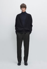 Cardigan nero in maglia con bottoni, abbinato a un dolcevita nero e a pantaloni scuri. L'outfit presenta una vestibilità comoda e una texture liscia.