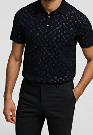 Homme portant un polo noir avec un motif de lettres bleues éparpillées et un pantalon noir, une main dans la poche, sur fond clair.
