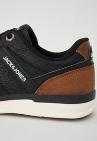 Black sneaker with textured synthetic upper, brown suede accent, cushioned collar, white midsole, and black rubber outsole with logo detail.