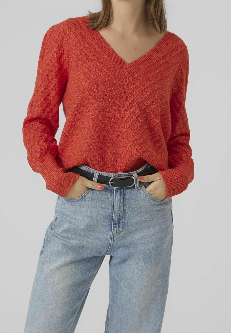 Femme portant un pull rouge à col en V texturé, rentré dans un jean bleu clair avec une ceinture noire, mains dans les poches, sur un fond uni.