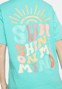 Tyrkis bomulls t-shirt med färgglad, lager-på-lager text som säger "SOLEN SKINER PÅ MINA TANKAR" med en solgrafik ovanför.