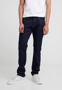 Person som bär mörkblå jeans, vita sneakers och en vit t-shirt som står mot en enfärgad ljus bakgrund.