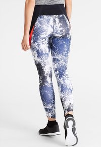 Kvinna som bär svart, vit och blå abstrakt mönstrade leggings med svart midjeband och svarta träningsskor, som går bort från en vit bakgrund.