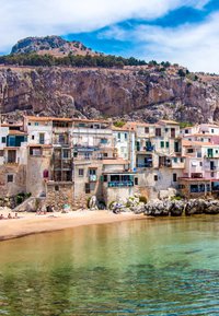 Küstengebäude mit weißen und pastellfarbenen Fassaden, einige mit Balkonen und grünen Fensterläden, in der Nähe eines sandigen Strandes und einer felsigen Küste unter blauem Himmel.