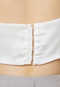 Top de cetim branco com um painel traseiro liso que apresenta um fecho metálico de gancho e olho; textura suave, design minimalista, sem padrões visíveis.