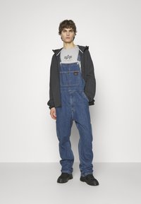 Jeansoveralls in Mittelblau mit verstellbaren Trägern, einer Vorder- und seitlichen Taschen, kombiniert mit einem grauen T-Shirt und schwarzen Schuhen.