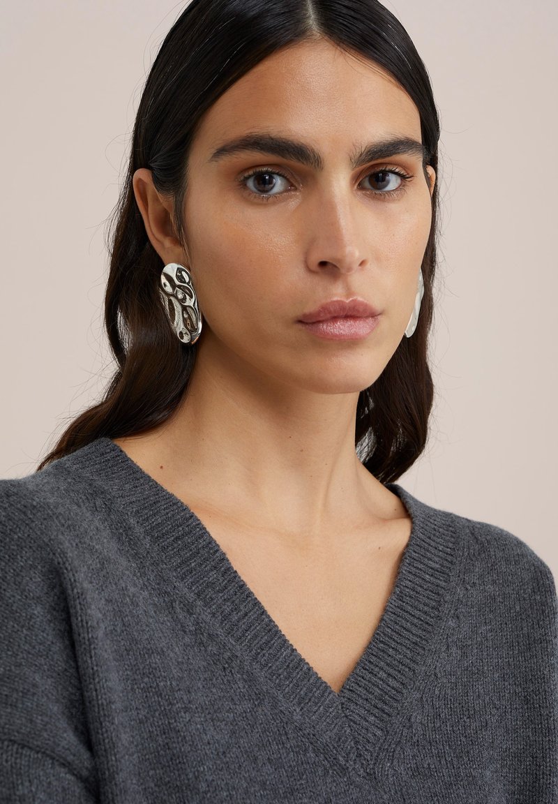 Grijze gebreide trui met een V-hals, gecombineerd met grote, organisch gevormde zilveren oorbellen met uitsnijdingen en gepolijste accenten.