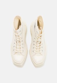 Høye sneakers i kremfarget lerret med glanset hvit gummitåhette, sølvfargede øyelettei og en svart logo-patch på utsiden.