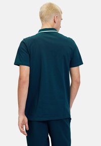 Polo shirt verde scuro con colletto e accenti bianchi. Maniche corte e texture liscia, mostrato da dietro con pantaloni di colore simile.
