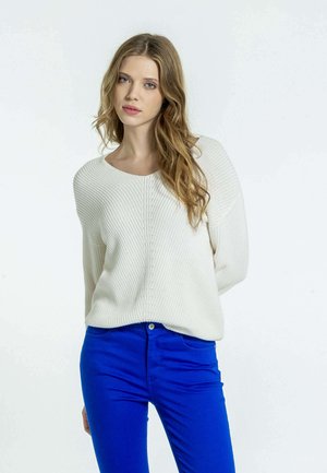 Pull en maille crème avec une texture côtelée, présentant un col large et des épaules tombantes, associé à un jean slim bleu vif.