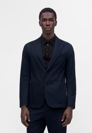Jeune homme portant une veste et un pantalon de costume bleu marine foncé avec une chemise noire boutonnée, debout devant un fond clair uni.