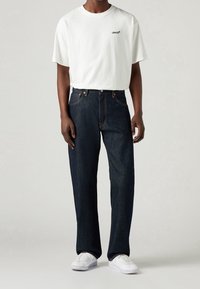 Vit bomullströja med liten svart "Levi's"-logga, kombinerad med mörkblå denimjeans och vita sneakers, med avslappnad passform och klassisk design.