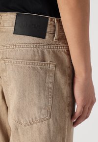 Beige denim jeans hebben een gestructureerd oppervlak, details op de achterzak en een zwart lederen logo patch met de tekst "ONLY & SONS."