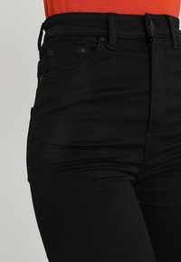 Jeans negros de tiro alto con un diseño ajustado, que cuentan con bolsillos delanteros y una textura suave. Los detalles de costura son visibles a lo largo de las costuras.