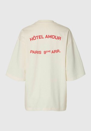 T-shirt oversize crème avec un texte rouge indiquant "HÔTEL AMOUR PARIS 9ÈME ARR." au dos, sur un fond uni.