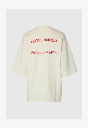 Krém színű bő fazonú póló, hátulján piros szöveg olvasható: "HÔTEL AMOUR PARIS 9EME ARR." egyszínű háttér előtt.