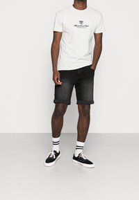 Camiseta de algodón gris con logo, pantalones cortos de mezclilla negra con puños enrollados y zapatillas negras con suelas blancas y calcetines de rayas.