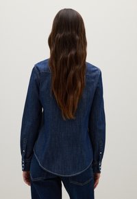 Woman with long wavy brown hair wearing a dark denim shirt and jeans, standing facing a plain light gray wall.