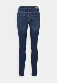 Jeans skinny em denim azul escuro com uma textura suave, apresentando dois bolsos traseiros e uma bainha desfiada. Etiqueta da marca visível na cintura.