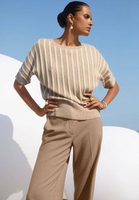 Vrouw in een beige gestreepte trui en wijde broek poseert buitenshuis met de handen op de heupen, tegen een heldere blauwe lucht en een witte gebogen structuur.