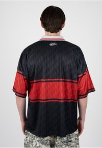 Hombre con camiseta de manga corta a rayas negras y rojas y pantalones de camuflaje beige, visto desde atrás sobre un fondo liso.