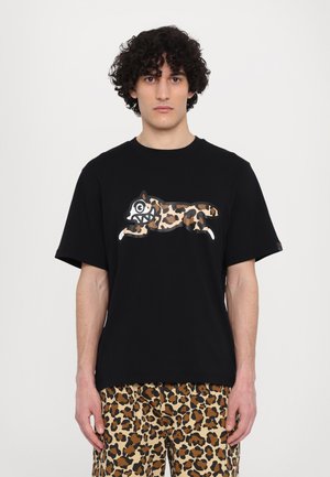 Schwarzes Baumwoll-T-Shirt mit kurzen Ärmeln, das auf der Vorderseite ein springendes Leopardenmotiv in Erdtönen zeigt. Das Model trägt gemusterte Leoparden-Shorts.