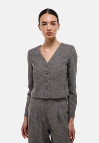 Blazer noir et blanc à motif pied de poule avec un col en V, des manches longues et cinq boutons. Tissu texturé avec une coupe ajustée.