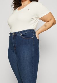 Weißes kurzärmliges V-Ausschnitt-Oberteil, kombiniert mit dunkelblauen hoch taillierten Jeans, die einen Frontknopf und ein dezentes Logo auf der Brust aufweisen.