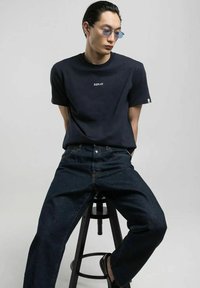 T-shirt in cotone navy con logo "REPLAY", abbinato a jeans in denim scuro. Il modello è seduto su uno sgabello nero, indossando occhiali da sole blu moderni.