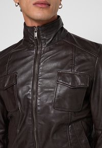 Braune Lederjacke mit hohem Kragen, Frontreißverschluss und zwei Brusttaschen. Sie verfügt über eine glatte Textur und kontrastierende Nahtdetails.