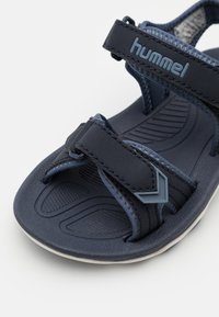 Mörkblå sandal med strukturerad gummisula, som har justerbara remmar och en framträdande logotyp. Inkluderar en dekorativ blå accent.