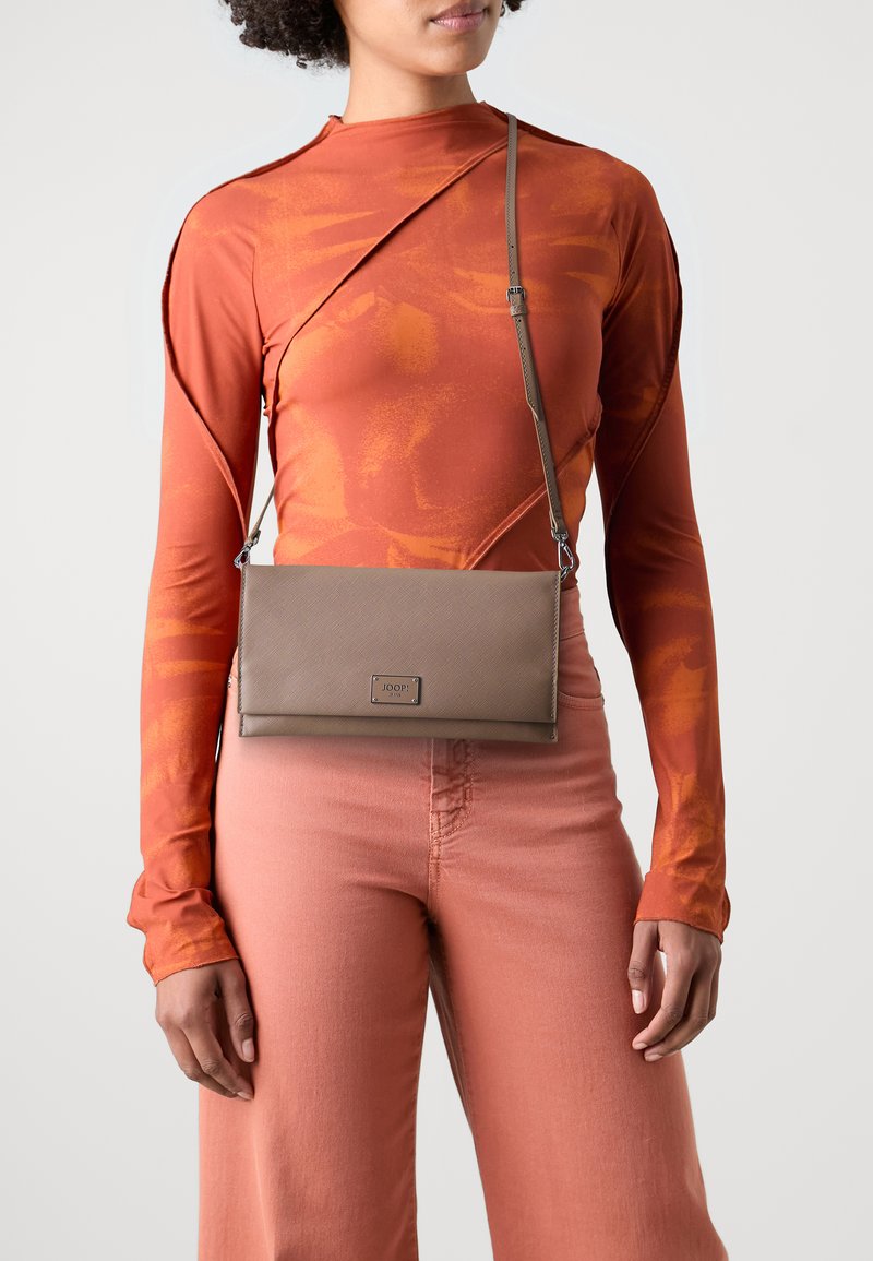 Hnědá texturovaná crossbody taška obdélníkového tvaru, se stříbrným kováním a logem značky, nošená s oranžovým dlouhým rukávem a růžovými kalhotami.