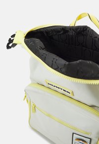 En ljusfärgad ryggsäck med gula accenter, som är utan dragkedja och avslöjar ett svart mönstrat inre samt flera externa fickor.