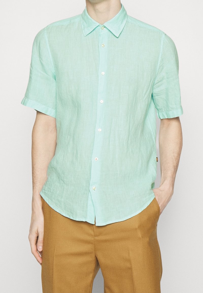 Jasno miętowa koszula z krótkim rękawem, wykonana z lnu, z guzikami z przodu i zaokrąglonym dołem. Połączona z beżowymi spodniami.