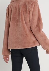 Rosa fuskpälskappa med krage, avslappnad passform och långa ärmar, sedd bakifrån, kombinerad med mörka jeans.