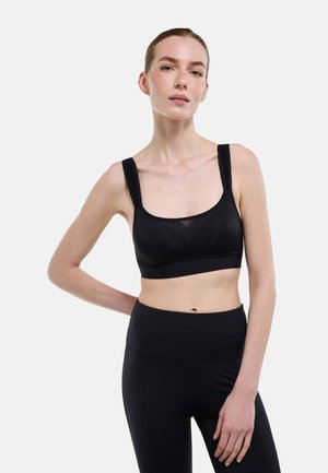 Femme portant un soutien-gorge de sport noir et un legging taille haute, debout avec une main sur la hanche devant un fond uni.
