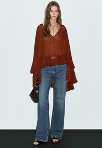 Blusa a maniche lunghe fluida color ruggine con orlo arricciato, abbinata a jeans svasati blu chiaro e una piccola borsa scura.