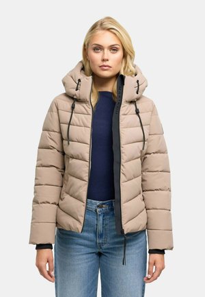 Blonde Frau trägt eine beige gesteppte Jacke mit Kapuze, dunkelblaues Oberteil und blaue Jeans und steht vor einem schlichten hellen Hintergrund.