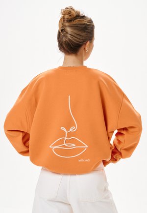 Vrouw met haar in een knot die een oranje sweatshirt draagt met een witte minimalistische lijntekening van een gezicht op de rug, gecombineerd met een witte broek.