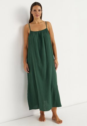 Kobieta stoi boso w luźnej, ciemnozielonej sukience do kostek z cienkimi ramiączkami na tle białej ściany.