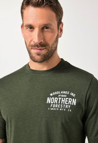 Tmavě zelené tričko s krátkým rukávem vyrobené z bavlněné směsi, s bílým tištěným textem a logem a dekorativními prvky na přední straně.