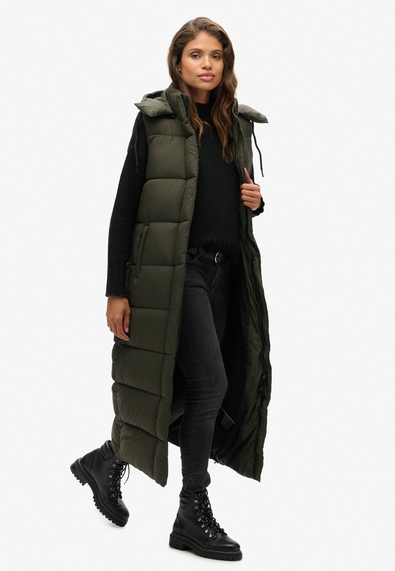 Langer, olivgrüner Steppweste mit Kapuze, mit einem Reißverschluss vorne und seitlichen Taschen, kombiniert mit einem schwarzen Pullover und grauen Jeans, abgerundet mit schwarzen Stiefeln.