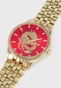 Gulv klokke med et rødt drage-design på en rød urskive. Har et guldfarvet metalarmbånd og en rillet bezel, med gyldne timemarkører og visere.